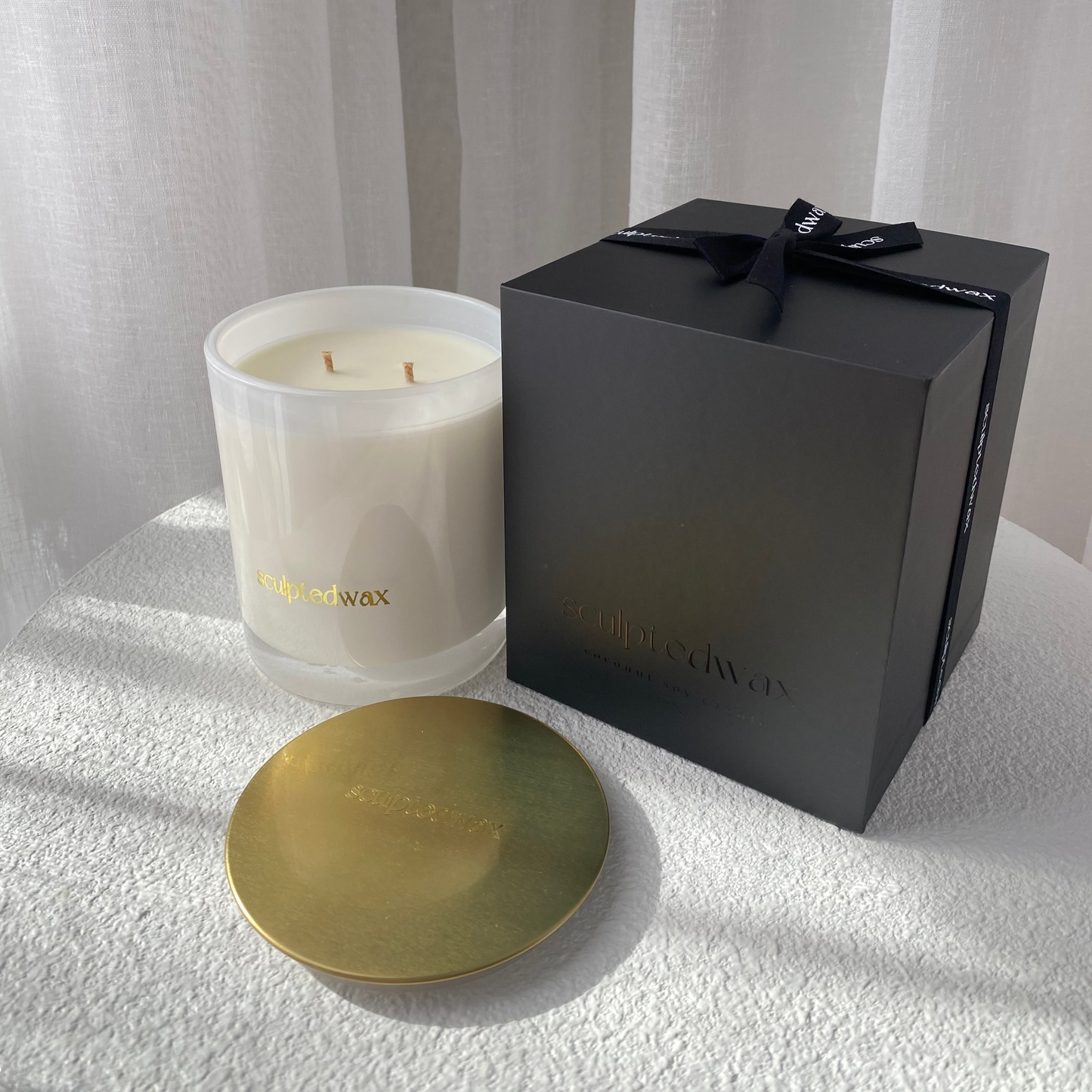 White candle with gold lid and black gift box on a white surface